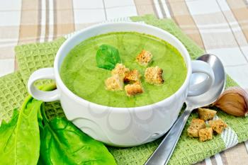 Green soup puree in a white bowl with croutons and spinach leaves, spoon on a napkin on the background fabric