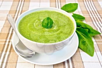 Cream soup green from spinach in a white bowl and saucer, spoon, spinach leaves on the background fabric