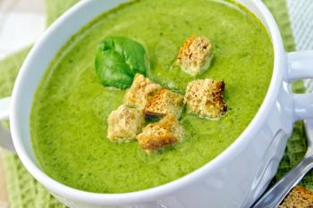 Green soup puree in a white bowl with croutons and spinach leaves, spoon on a napkin on the background fabric