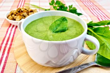Green soup puree in a bowl with green leaf spinach, spoon on the board, croutons, parsley on a cloth background