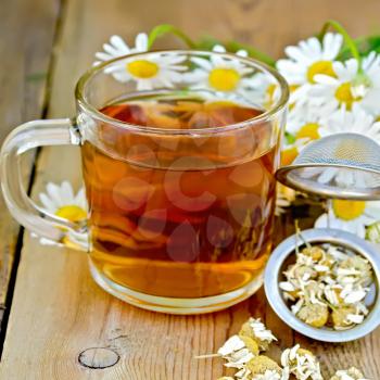 Tea in glass mug, metal sieve with dry chamomile flowers, a bouquet of fresh flowers daisies on a background of wooden boards