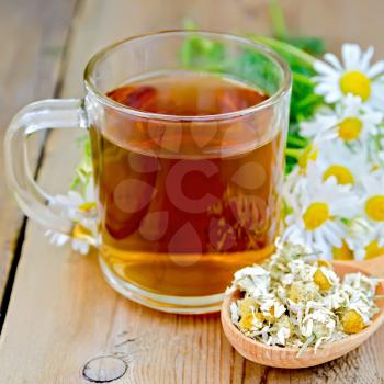 Glass mug with tea, wooden spoon with dry chamomile flowers, a bouquet of fresh chamomile flowers on a background of wooden boards