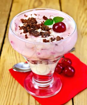 Milk dessert with cherry, chocolate biscuit and curd, a spoon, a red paper napkin, cherries on a background of wooden boards