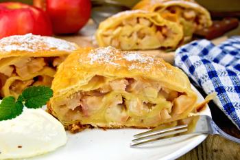 Strudel with apples and ice cream in a white plate with a fork, apples, knife, napkin on the background of wooden boards