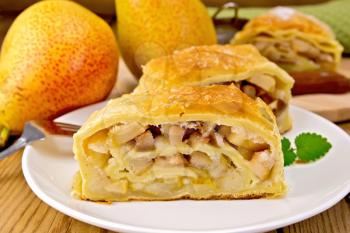 Strudel with pears in a white plate with a fork, pear, knife, napkin on the background of wooden boards