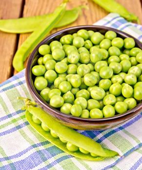 Green peas in a brown bowl on a checkered napkin, pea pods on the background of wooden boards
