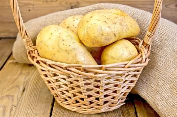 Tubers of yellow potatoes in a wicker basket on a sacking on a wooden boards background
