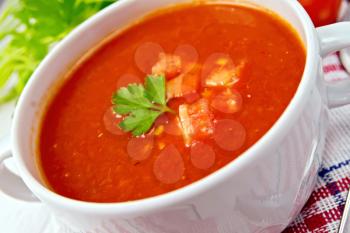 Tomato soup with chunks of vegetables in a white bowl on a napkin with a spoon, tomatoes, parsley on a lighter background board