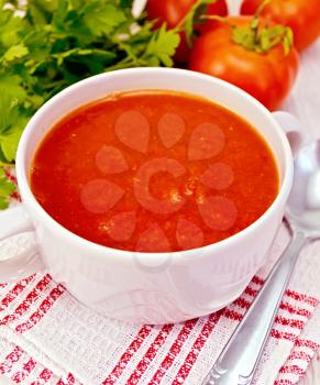 Tomato soup in a white bowl on a napkin with a spoon, tomatoes, parsley on a lighter background board