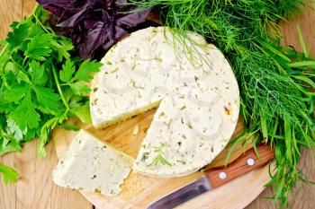 Homemade round cheese with herbs and spices, knife, parsley, basil, dill, tarragon, rosemary on a wooden boards background