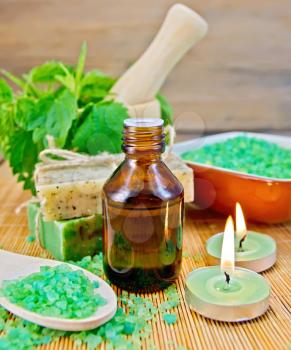 Oil in a bottle, two bars of homemade soap, bath salt, two scented candles, nettles in a mortar on a wooden boards background
