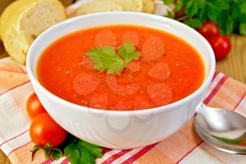 Tomato soup in a white bowl on a napkin, spoon, tomatoes, parsley, bread on a wooden boards background