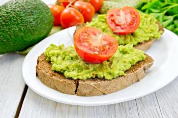 Two slices of rye bread with guakomole and tomato on a plate, napkin, parsley, avocado on a background of light wooden boards