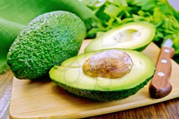 One whole and one halved avocado with bone, knife, parsley, green cloth on a wooden boards background