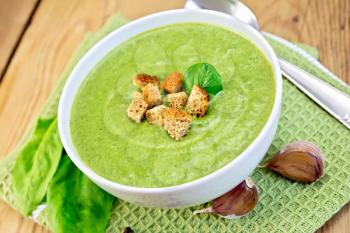 Green soup puree in a bowl with green spinach leaves, spoon, garlic on a napkin on a wooden boards background