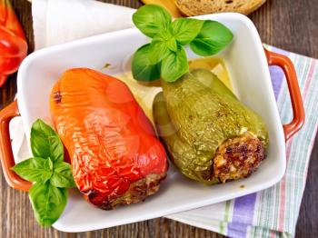Two sweet peppers stuffed with meat and rice with basil leaves in a brown roasting pan on a napkin on a wooden boards background on top