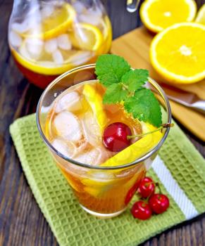 Lemonade in a glass and a jug with cherry, lemon and orange, mint green napkin on board with fruit on the background of dark wooden board