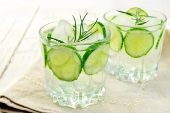 Lemonade with a cucumber and rosemary in two glassful on beige napkin against the background light wooden boards