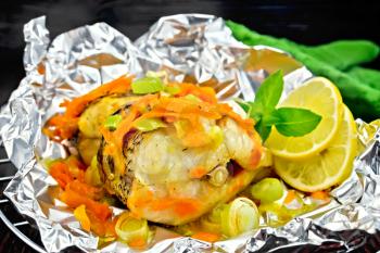 Pike with carrots, leek, basil and lemon slices in a foil on a metal grid, green towel on a dark wooden board