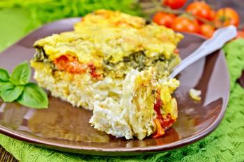 The pie of potatoes, cheese, tomato and spinach, filled egg with milk in a plate with a fork on a green napkin on a wooden boards background