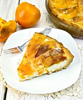 Sweet pie with curd and persimmons, a fork in the white plate on a napkin silicone, glass pan with pie on the background light wooden boards