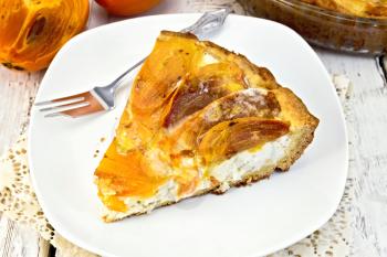 Sweet pie with curd and persimmons in a white plate, a fork in the white plate on a napkin openwork silicon, glass pan with pie on a wooden boards background