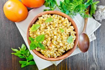 Barley porridge in a clay bowl with basil on a napkin, tomatoes, parsley and garlic on a background of wooden boards on top