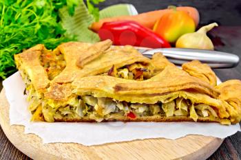 Pie with cabbage, sorrel and sweet pepper on parchment, knife, onion and parsley on a wooden board background