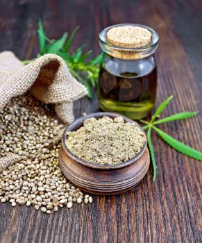 Hemp flour in a clay bowl, the grain in the bag and on the table, the oil in a glass jar, cannabis leaves on the background of wood planks