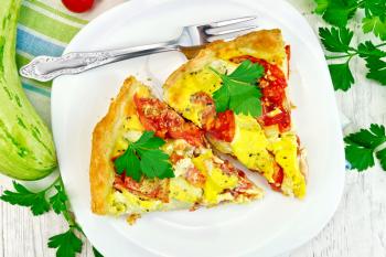Two pieces pie of zucchini with tomatoes and eggs in a plate on a napkin, parsley on a background of wooden boards on top