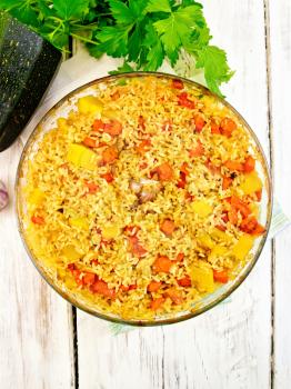 Rice with chicken, tomatoes, carrots, zucchini and garlic in a glass pan on a napkin, parsley on a background of wooden boards on top