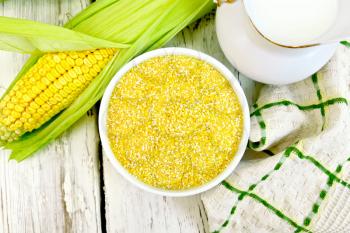 Corn grits in a white bowl, cobs, a jug of milk and a napkin on a background of wooden boards on top