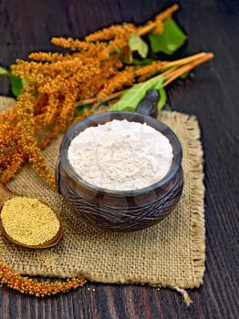 Flour amaranth in a clay cup, a spoon with grain, brown flower with leaves on a napkin from a sacking on a background of wooden boards