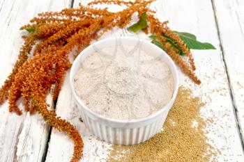 Amaranth flour in a bowl, seeds scattered on the table, brown flower with green leaves on a background of wooden boards