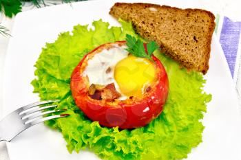 Scrambled eggs with ham and mushrooms in a tomato on a green lettuce in the plate, bread, fork and a napkin on the background light wooden boards