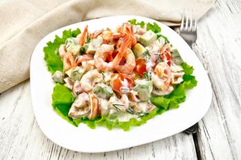 Salad with shrimp, avocado, tomatoes and mayonnaise on the green lettuce in a white plate, napkin, fork on the background light wooden table