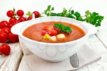 Gazpacho tomato soup in a white bowl with parsley and vegetables, spoon on a napkin against the background of wooden boards