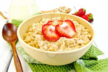 Oatmeal porridge in a bamboo bowl with strawberries on a green napkin, spoon, oat flakes in a spoon on a light wooden board background