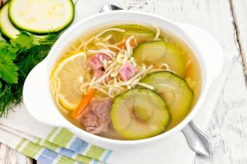 Soup with zucchini, beef, ham, lemon and noodles in a bowl on a napkin, parsley and dill on a wooden plank background