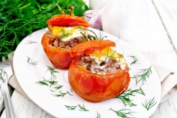 Tomatoes stuffed with meat and rice with cheese in a plate on a napkin, fork, dill and parsley on a wooden board background