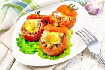 Tomatoes stuffed with meat and rice with cheese on lettuce in a plate, towel, fork against a light wooden board
