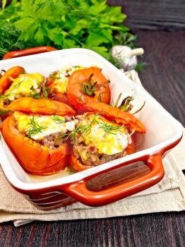 Tomatoes stuffed with meat and rice with cheese in a brazier on a napkin, parsley, dill and garlic on a wooden board background