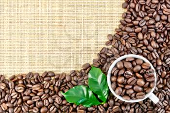 A frame of black coffee beans with green leaves and a cup on a yellow coarse woven fabric