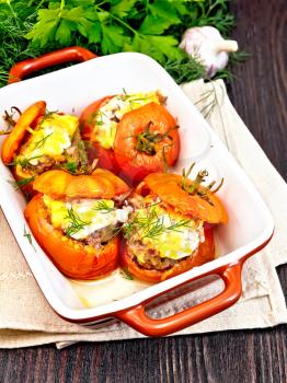 Tomatoes stuffed with meat and rice with cheese in a brazier on a light napkin, parsley, dill and garlic on a wooden board background