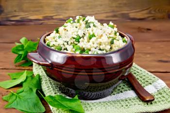 Couscous with spinach and green peas in a clay bowl on a napkin, basil and spoon on a wooden plank background