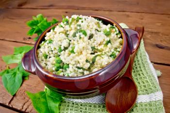Couscous with spinach and green peas in a clay bowl on a kitchen towel, basil and spoon on a wooden plank background