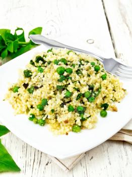 Couscous with spinach and green peas in a plate on a napkin, basil and fork on a light wooden board background