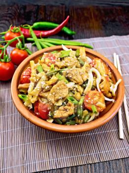 Thai noodles wok with chicken meat, tomatoes, soy sauce and green beans sprinkled with sesame seeds in a plate on a bamboo napkin on a wooden board background
