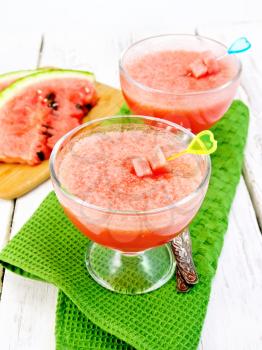 Jelly airy watermelon in two glass bowls, a spoon on a green towel on a wooden plank background