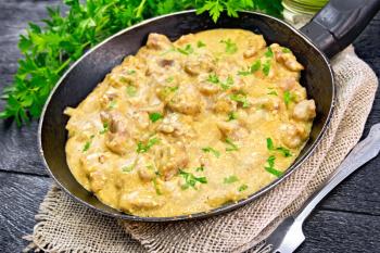 Meat stewed with cream in an old frying pan on burlap, parsley, fork on a wooden plank background